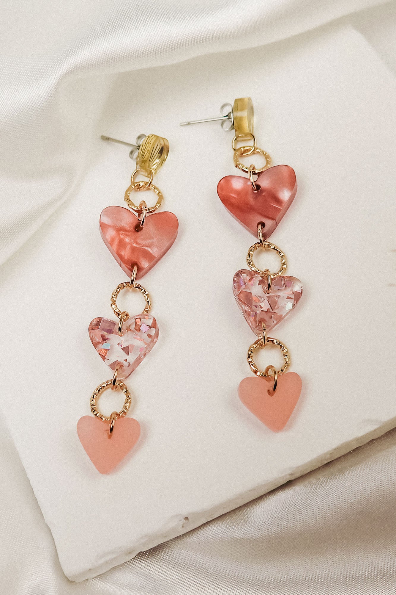 A pair of Heart Chain Earrings featuring three heart-shaped charms in pink tones, linked with gold rings for allergy-free comfort, displayed on a white textured surface with a satin fabric background.
