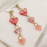 A pair of Heart Chain Earrings featuring three linked heart-shaped charms in red, pink, and light pink tones with gold rings, designed for allergy-free comfort and displayed on a soft white fabric background.