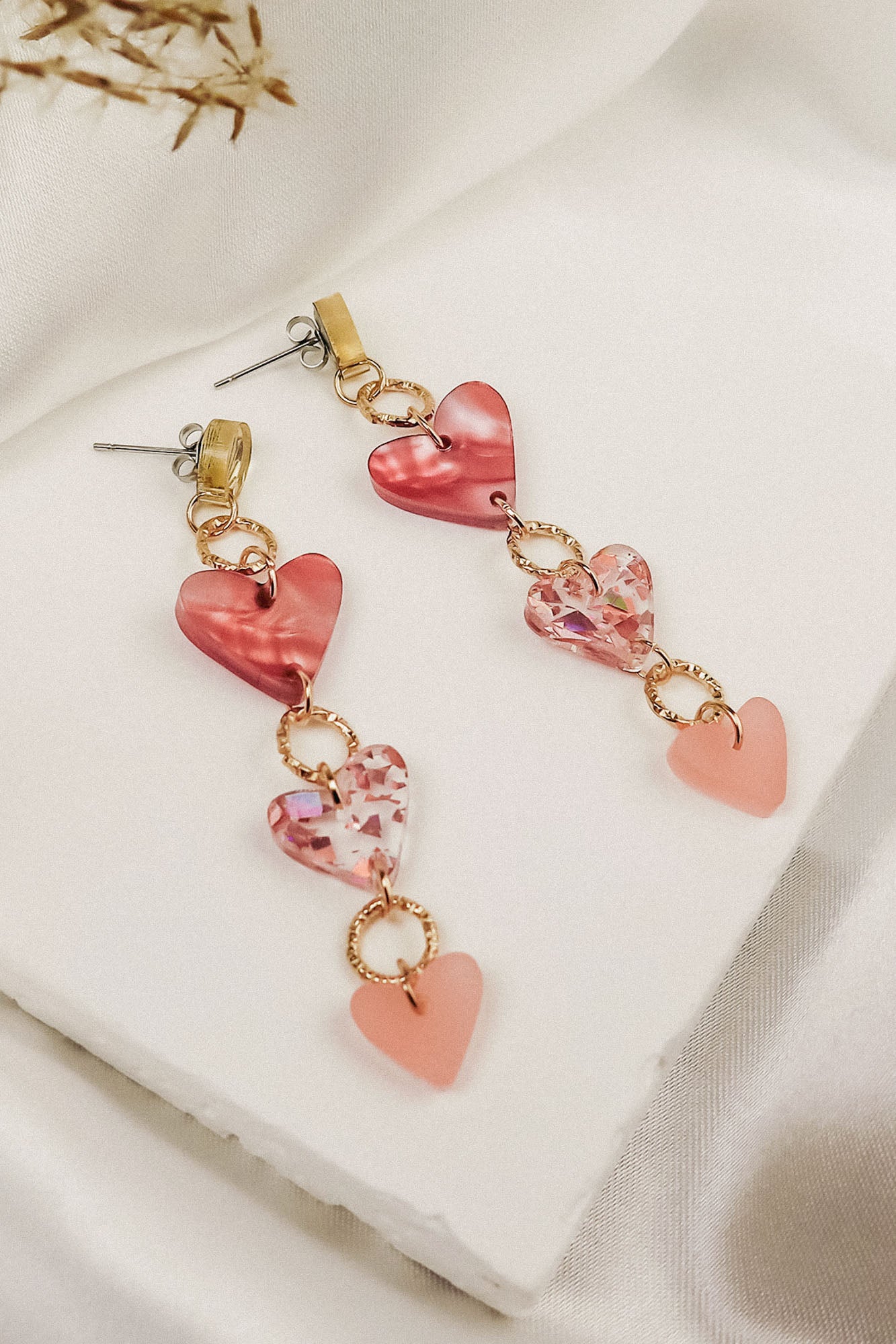 A pair of Heart Chain Earrings featuring three linked heart-shaped charms in red, pink, and light pink tones with gold rings, designed for allergy-free comfort and displayed on a soft white fabric background.