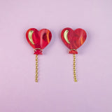 Two red marbled Heart Balloon Studs with small gold crescent details and playful gold chains hanging from the bottom, placed side by side on a light purple background.