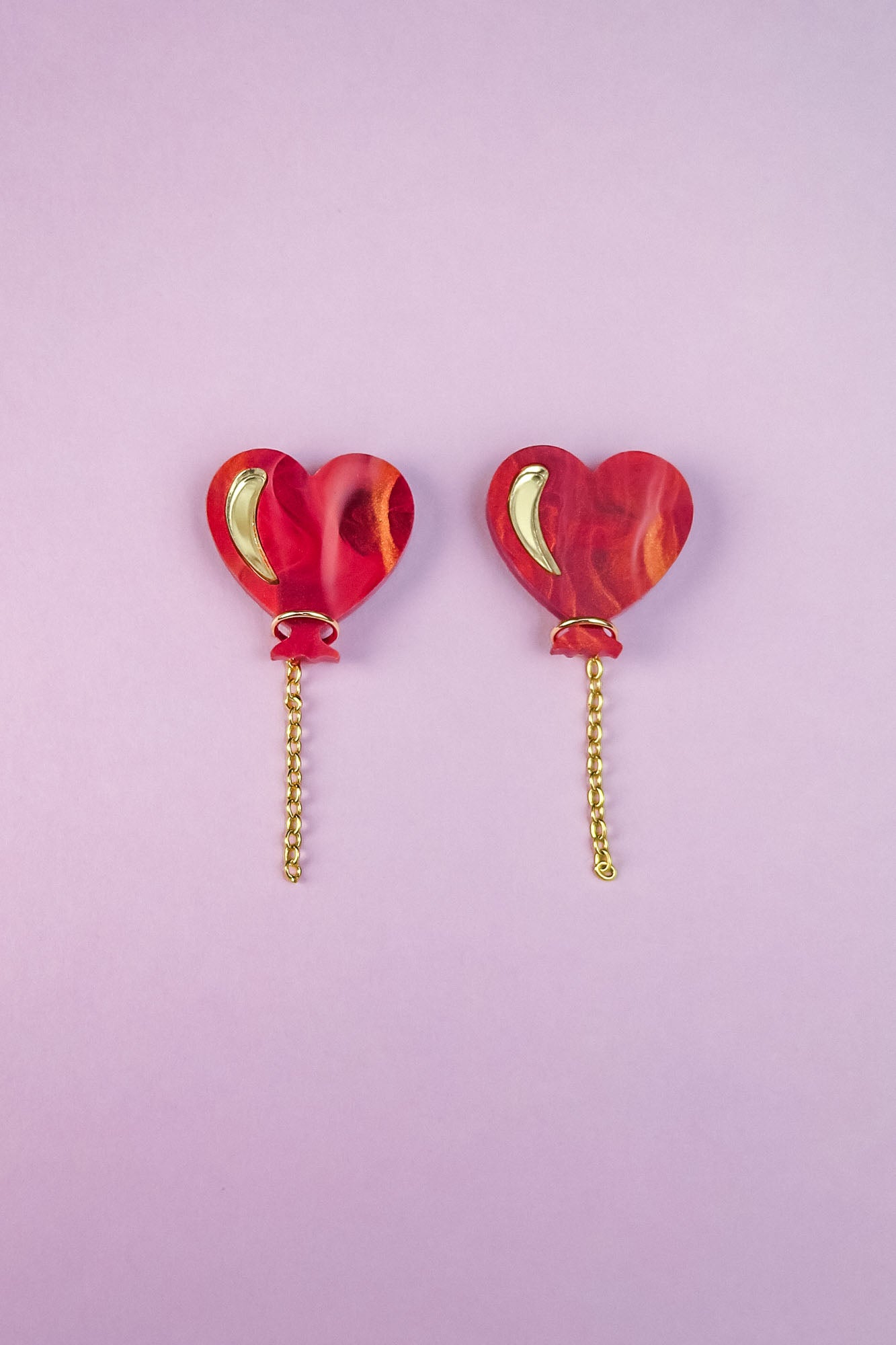 Two red marbled Heart Balloon Studs with small gold crescent details and playful gold chains hanging from the bottom, placed side by side on a light purple background.