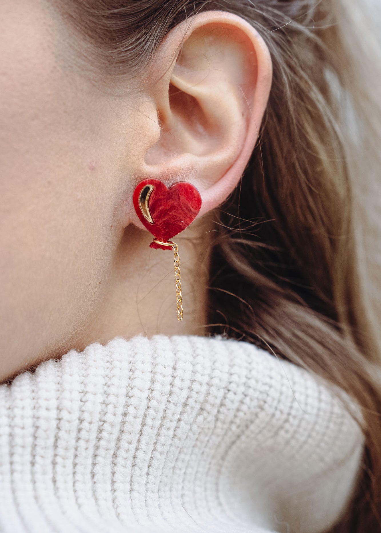 Close-up of a woman's ear wearing playful Heart Balloon Studs—red heart-shaped earrings with a small gold chain. Part of her face and blonde hair is visible, complemented by a white ribbed turtleneck sweater.