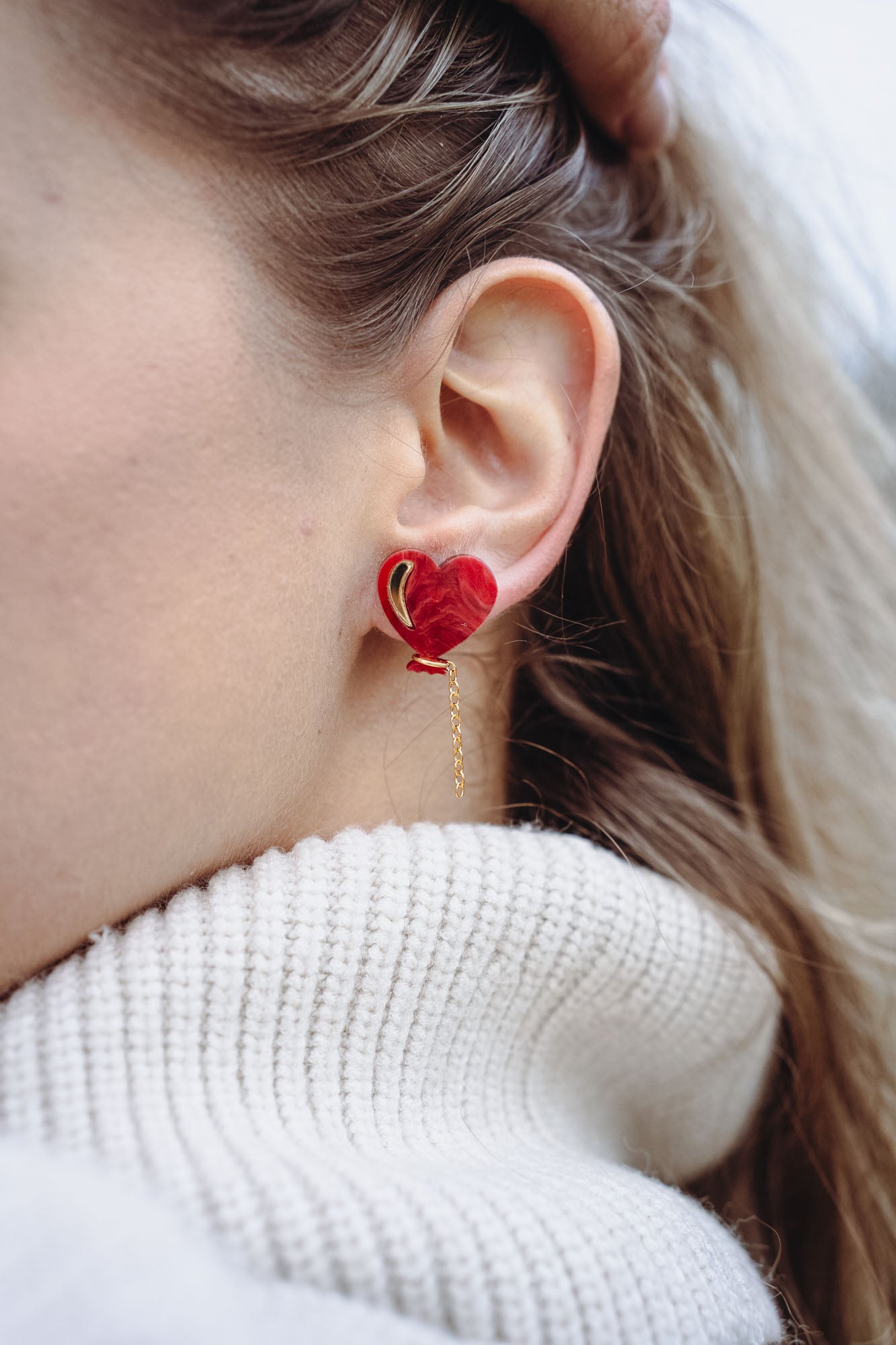 Close-up of a woman's ear wearing playful Heart Balloon Studs—red heart-shaped earrings with a small gold chain. Part of her face and blonde hair is visible, complemented by a white ribbed turtleneck sweater.