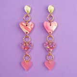 A pair of Heart Chain Earrings with gold studs and three pink and purple heart-shaped charms hanging from each, crafted for allergy-free comfort, displayed on a purple background.