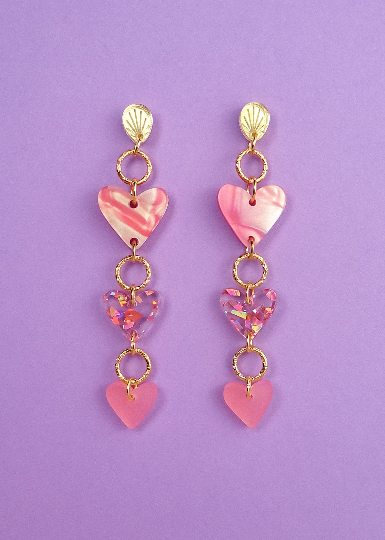 A pair of Heart Chain Earrings with gold studs and three pink and purple heart-shaped charms hanging from each, crafted for allergy-free comfort, displayed on a purple background.