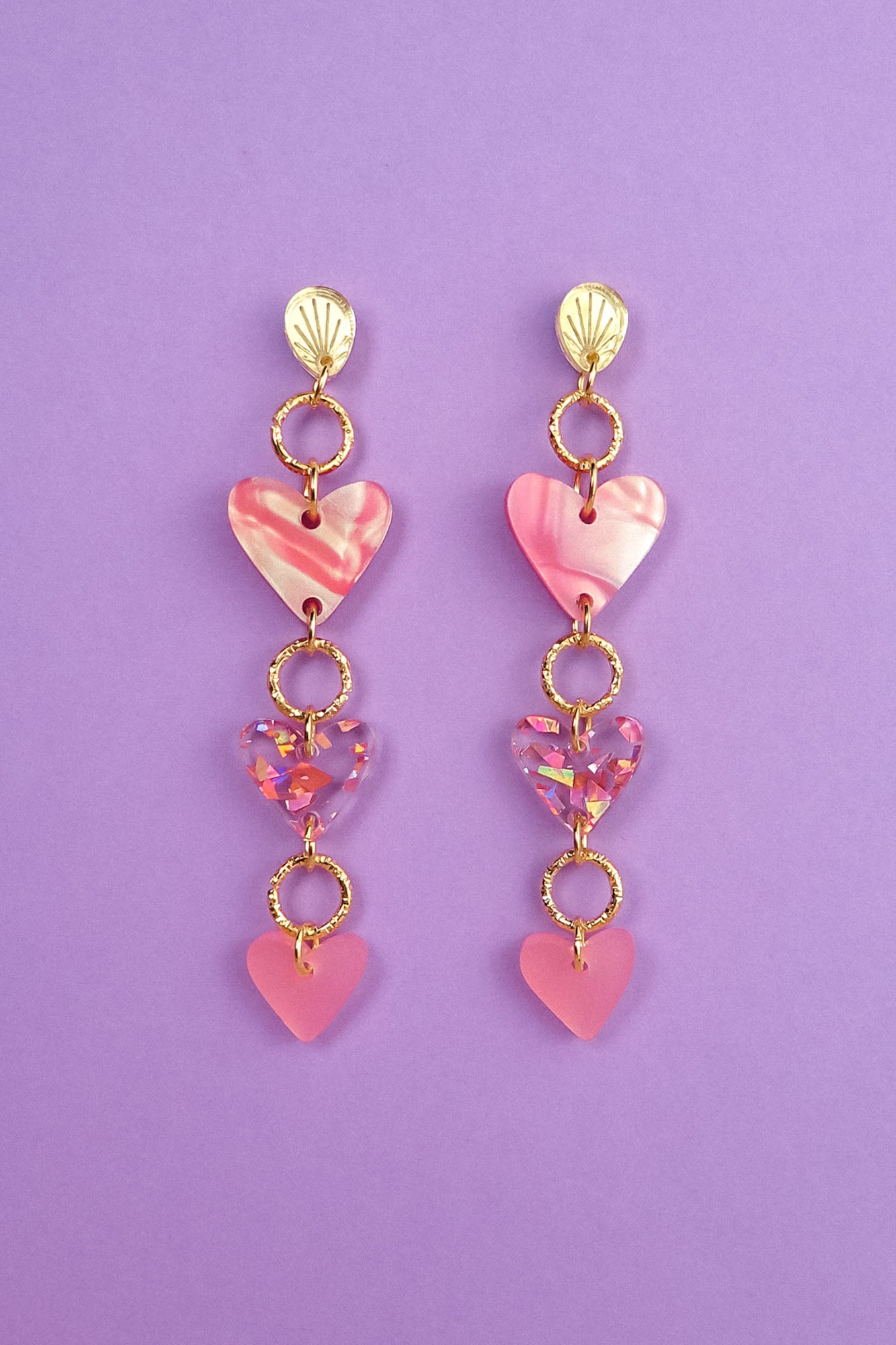 A pair of Heart Chain Earrings with gold studs and three pink and purple heart-shaped charms hanging from each, crafted for allergy-free comfort, displayed on a purple background.