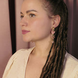 A woman with long brown dreadlocks and gold Heart Chain Earrings gazes to the left. She wears a cream-colored blouse and stands against a backdrop of pink curtains, highlighting soft pink tones throughout the scene.