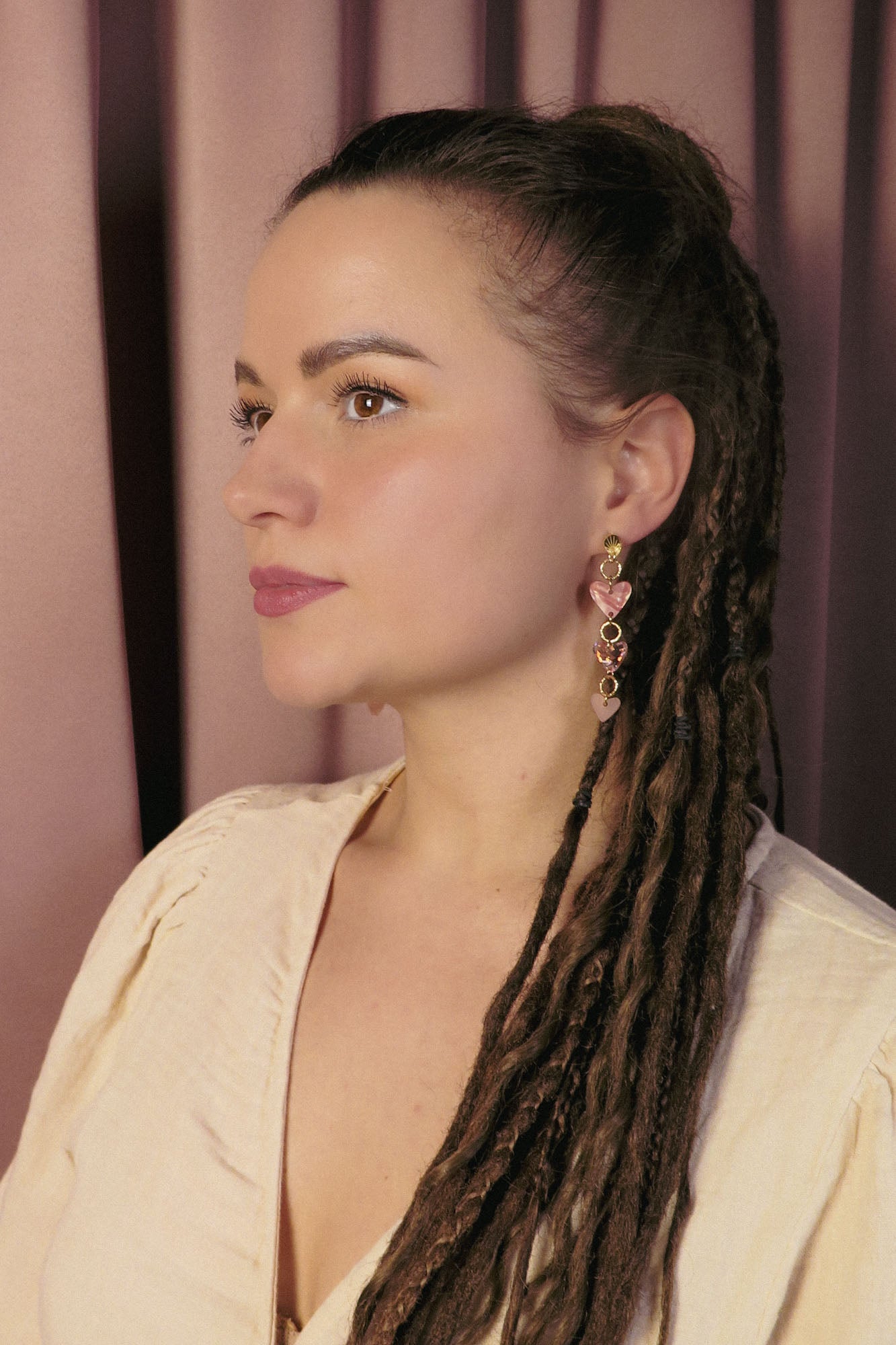 A woman with long brown dreadlocks and gold Heart Chain Earrings gazes to the left. She wears a cream-colored blouse and stands against a backdrop of pink curtains, highlighting soft pink tones throughout the scene.