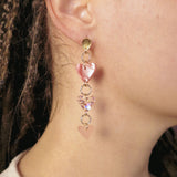 Close-up of a person wearing Heart Chain Earrings in gold and pink tones. They have braided hair and a light-colored top, with an out-of-focus background. Designed for allergy-free comfort, these earrings add a playful touch to any look.