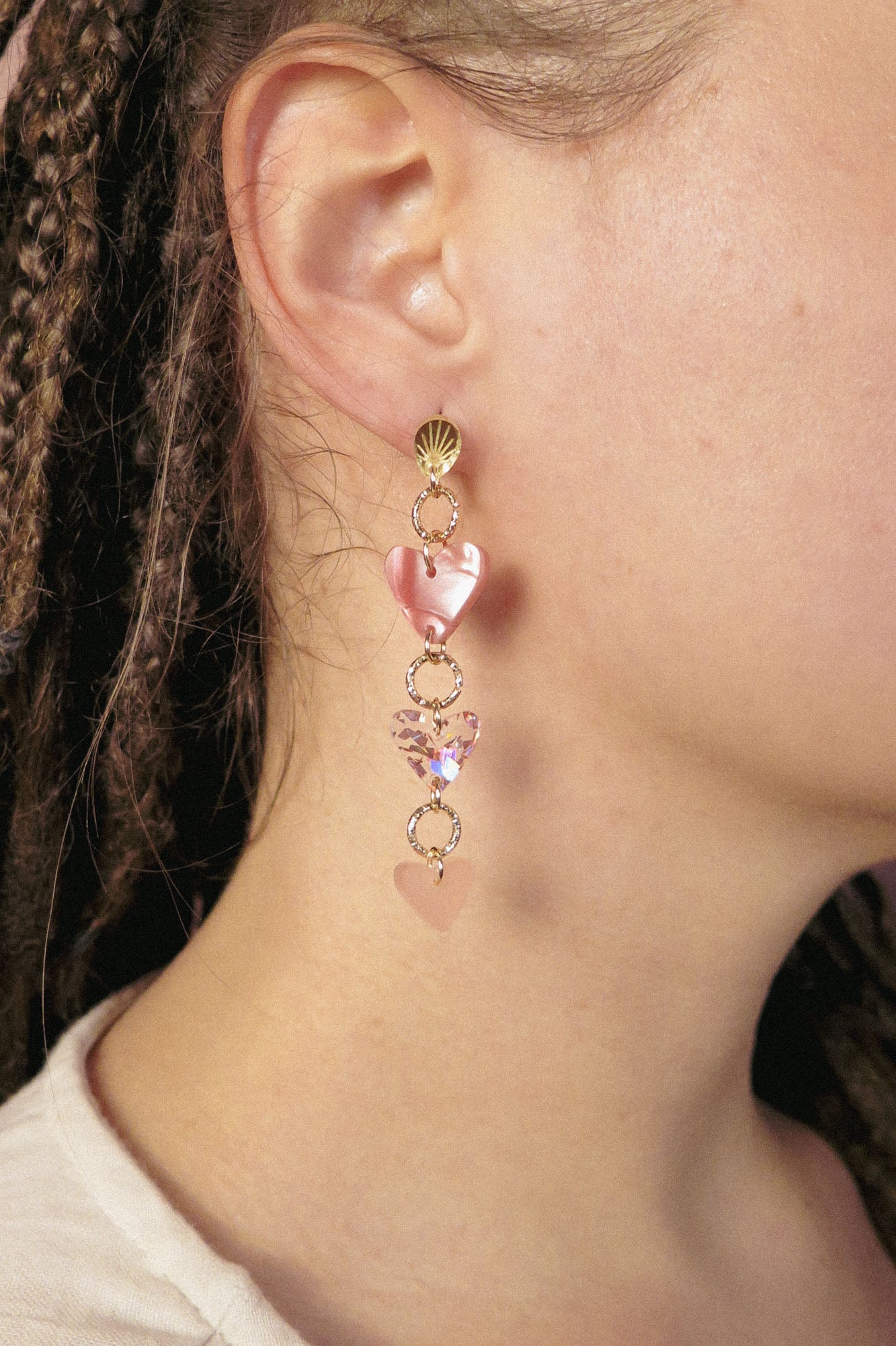 Close-up of a person wearing Heart Chain Earrings in gold and pink tones. They have braided hair and a light-colored top, with an out-of-focus background. Designed for allergy-free comfort, these earrings add a playful touch to any look.