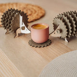 Two wooden hedgehog-shaped felt coasters hold a round, pink cup on a wooden table with woven placemats in the background—a charming touch of home decor.