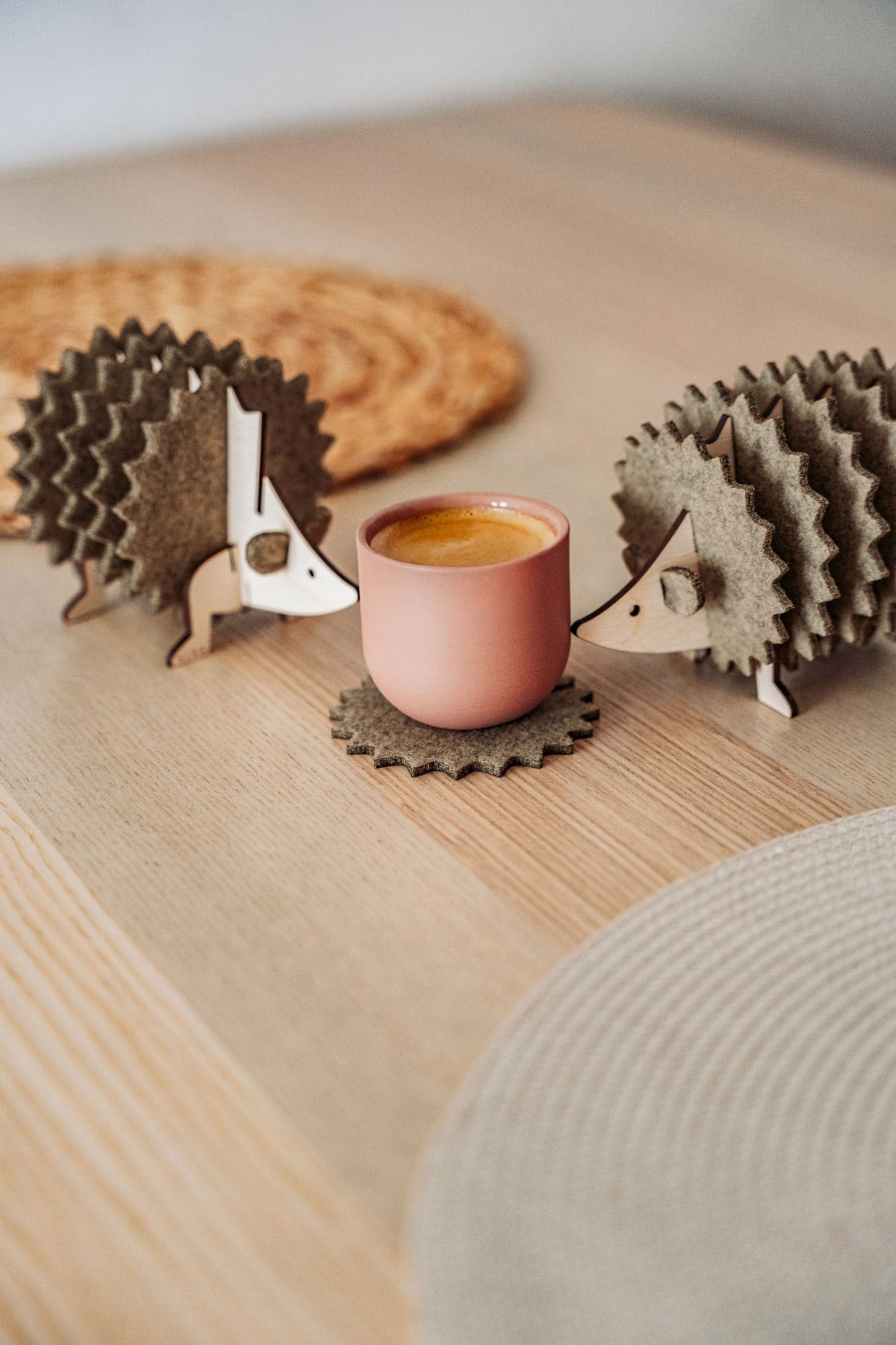 Two wooden hedgehog-shaped felt coasters hold a round, pink cup on a wooden table with woven placemats in the background—a charming touch of home decor.