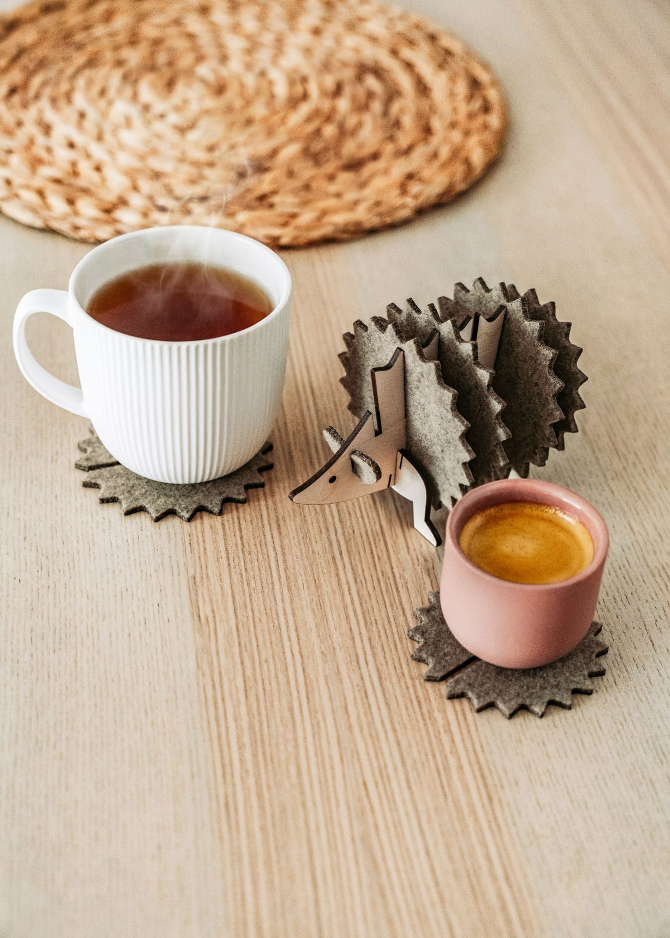 A white mug and a pink cup sit on spiky felt coasters shaped like a hedgehog, adding playful home decor flair to a light wooden table with a woven placemat in the background.