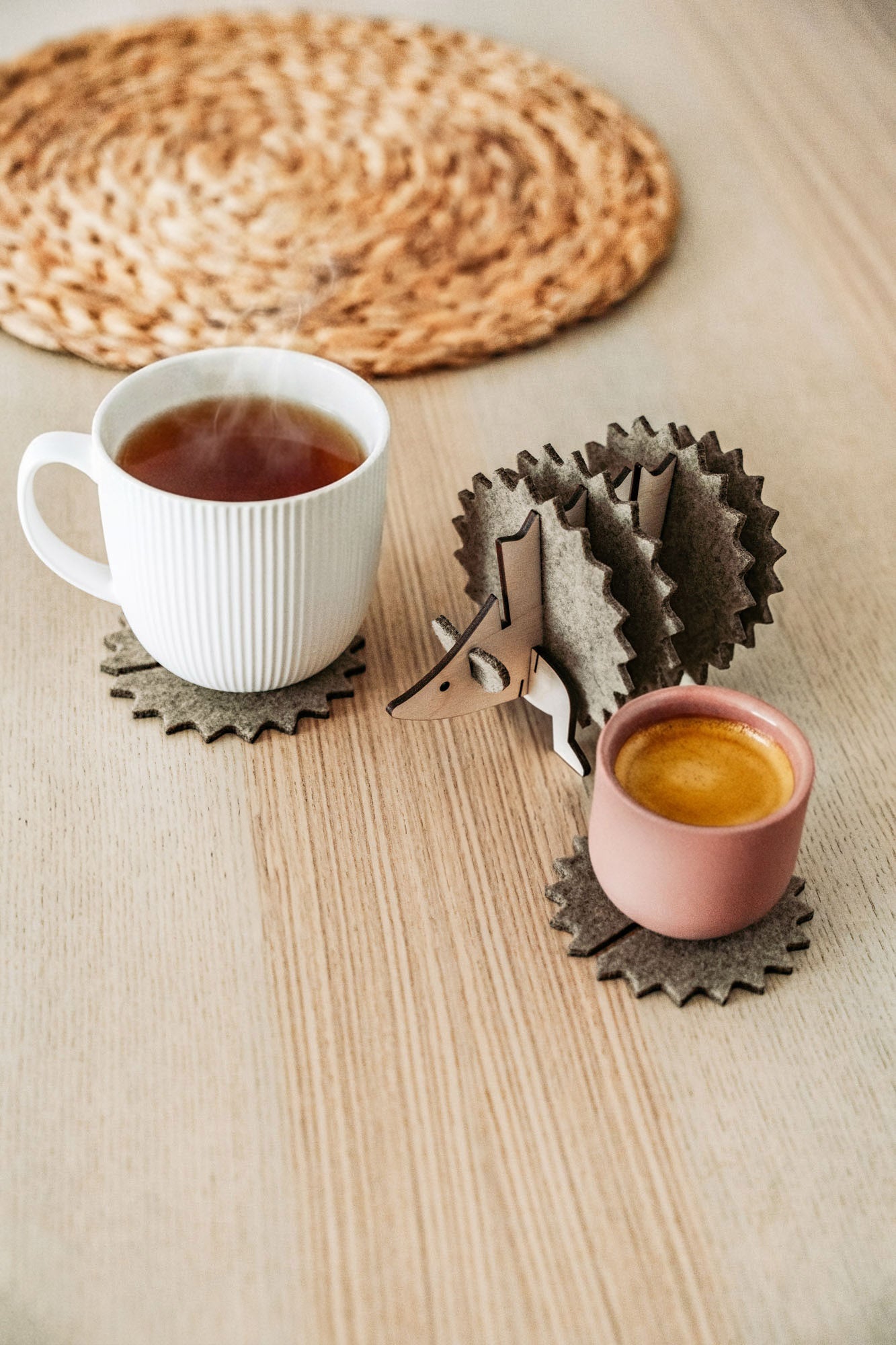 A white mug and a pink cup sit on spiky felt coasters shaped like a hedgehog, adding playful home decor flair to a light wooden table with a woven placemat in the background.