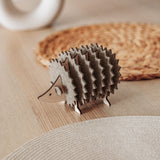 A small, decorative wooden hedgehog coaster holder sits on a wooden table with a woven placemat and a round, white ceramic object in the background, adding charm to your home decor.