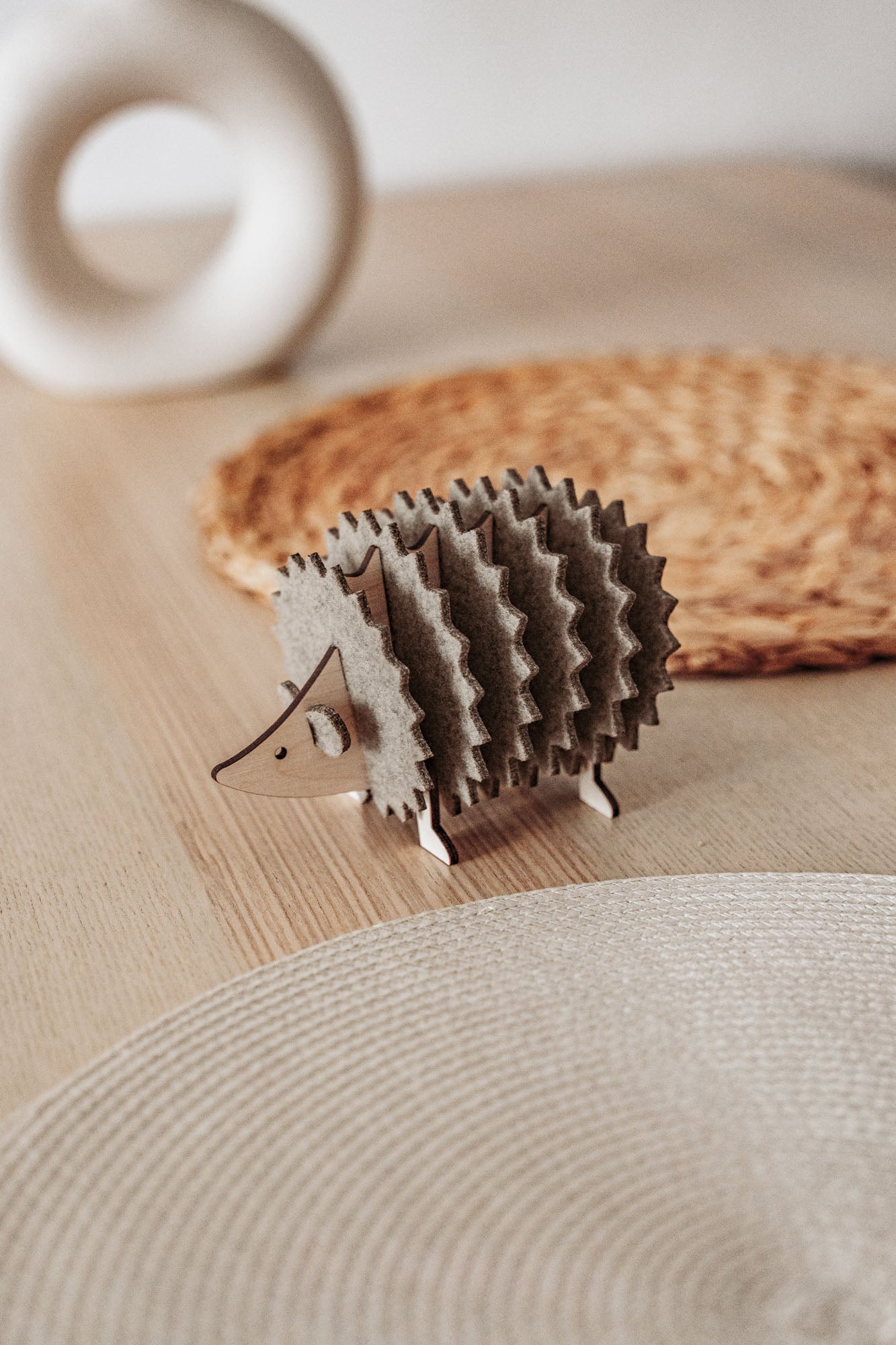 A small, decorative wooden hedgehog coaster holder sits on a wooden table with a woven placemat and a round, white ceramic object in the background, adding charm to your home decor.