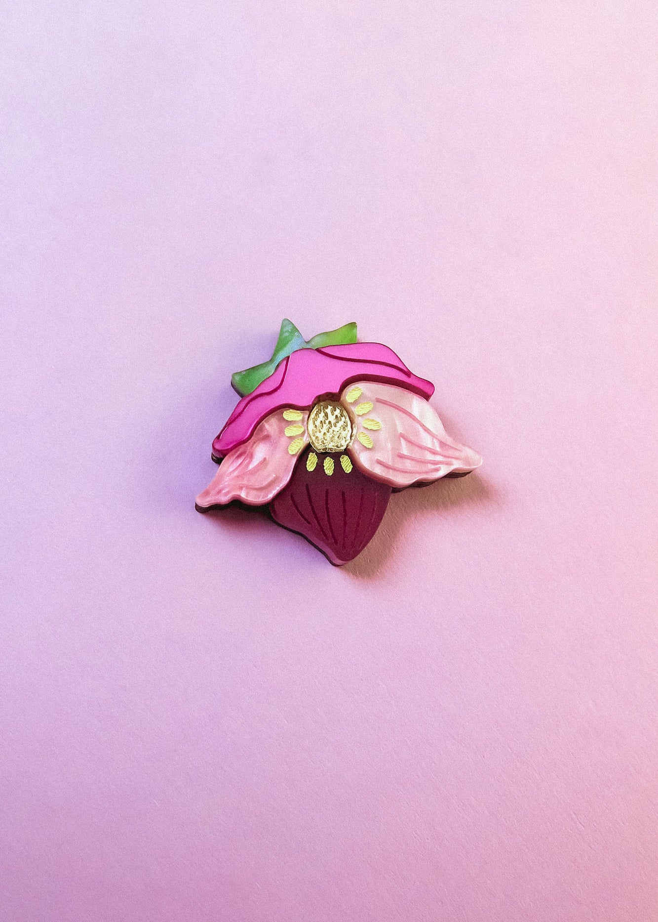 A colorful enamel lapel pin shaped like a strawberry with pink petals and green leaves is displayed on a soft pink background—perfect for handmade accessory lovers or fans of the unique purple hellebore flower.