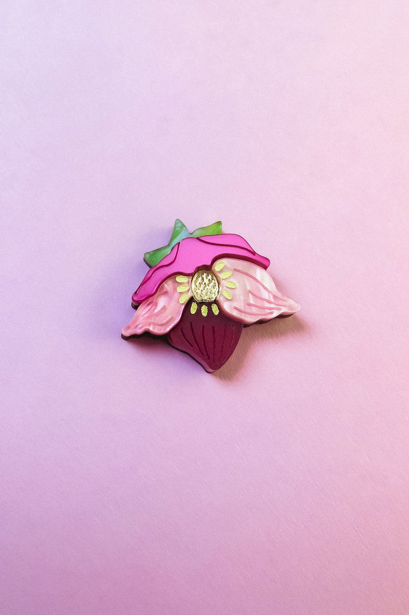 A colorful enamel lapel pin shaped like a strawberry with pink petals and green leaves is displayed on a soft pink background—perfect for handmade accessory lovers or fans of the unique purple hellebore flower.