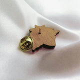 A handmade wooden lapel pin shaped like a maple leaf with a gold-colored clasp is resting on smooth white fabric.