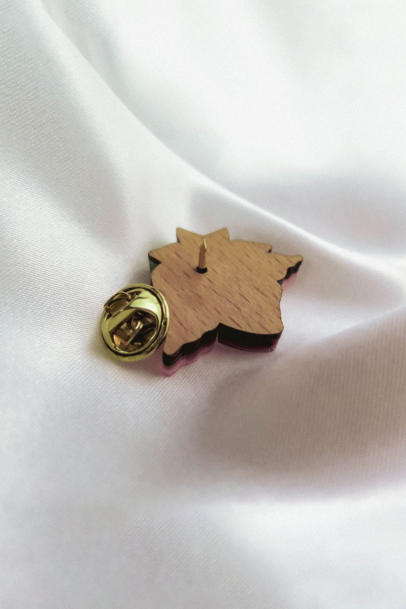 A handmade wooden lapel pin shaped like a maple leaf with a gold-colored clasp is resting on smooth white fabric.