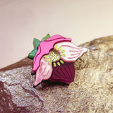 A colorful handmade lapel pin shaped like a purple hellebore flower, with pink petals, a green stem, and a yellow center, rests on a textured, metallic rock surface.