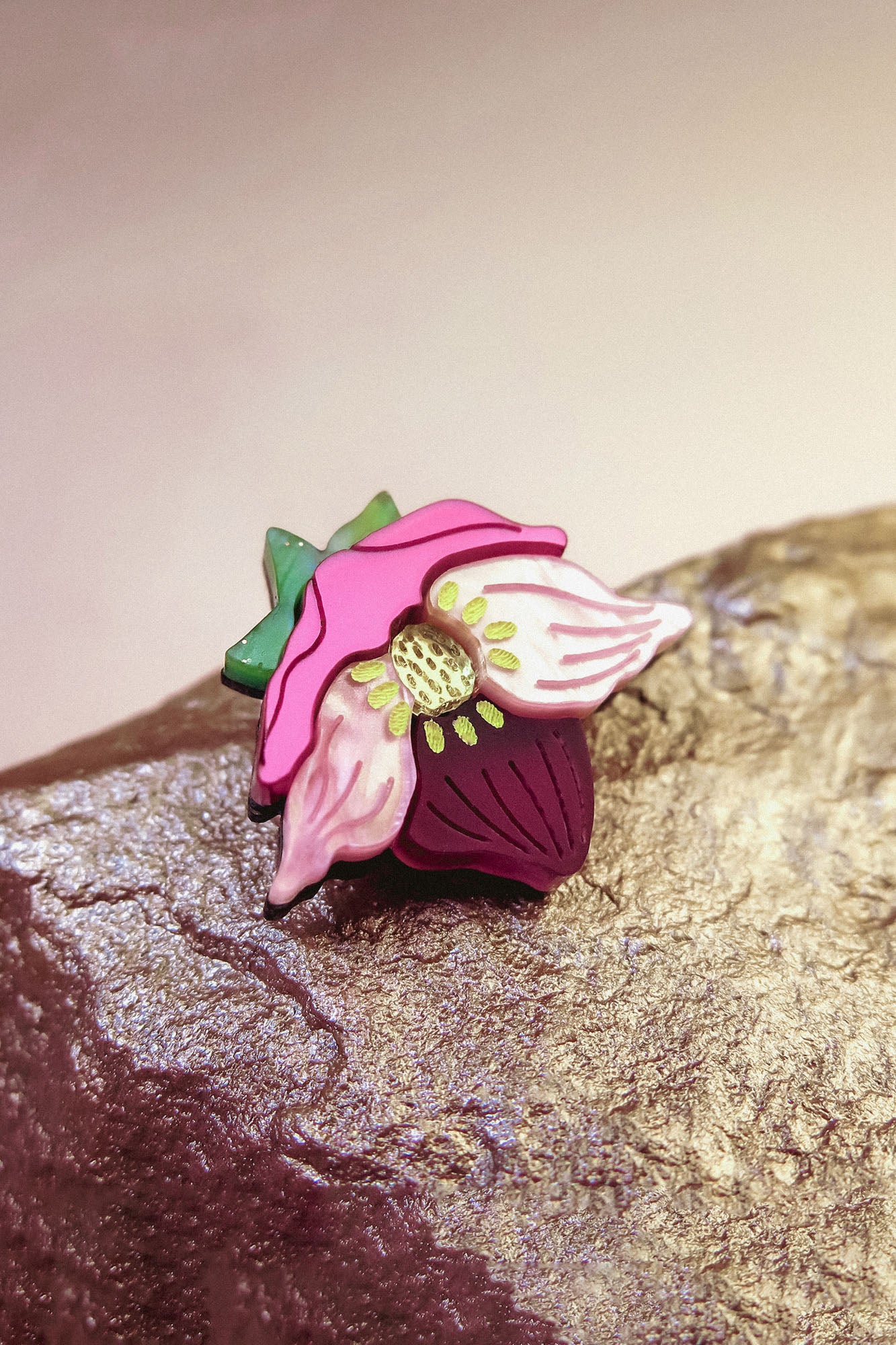 A colorful handmade lapel pin shaped like a purple hellebore flower, with pink petals, a green stem, and a yellow center, rests on a textured, metallic rock surface.