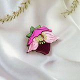 A handmade Purple Hellebore Flower lapel pin rests on white satin fabric, with delicate sprigs of dried grass in the background.