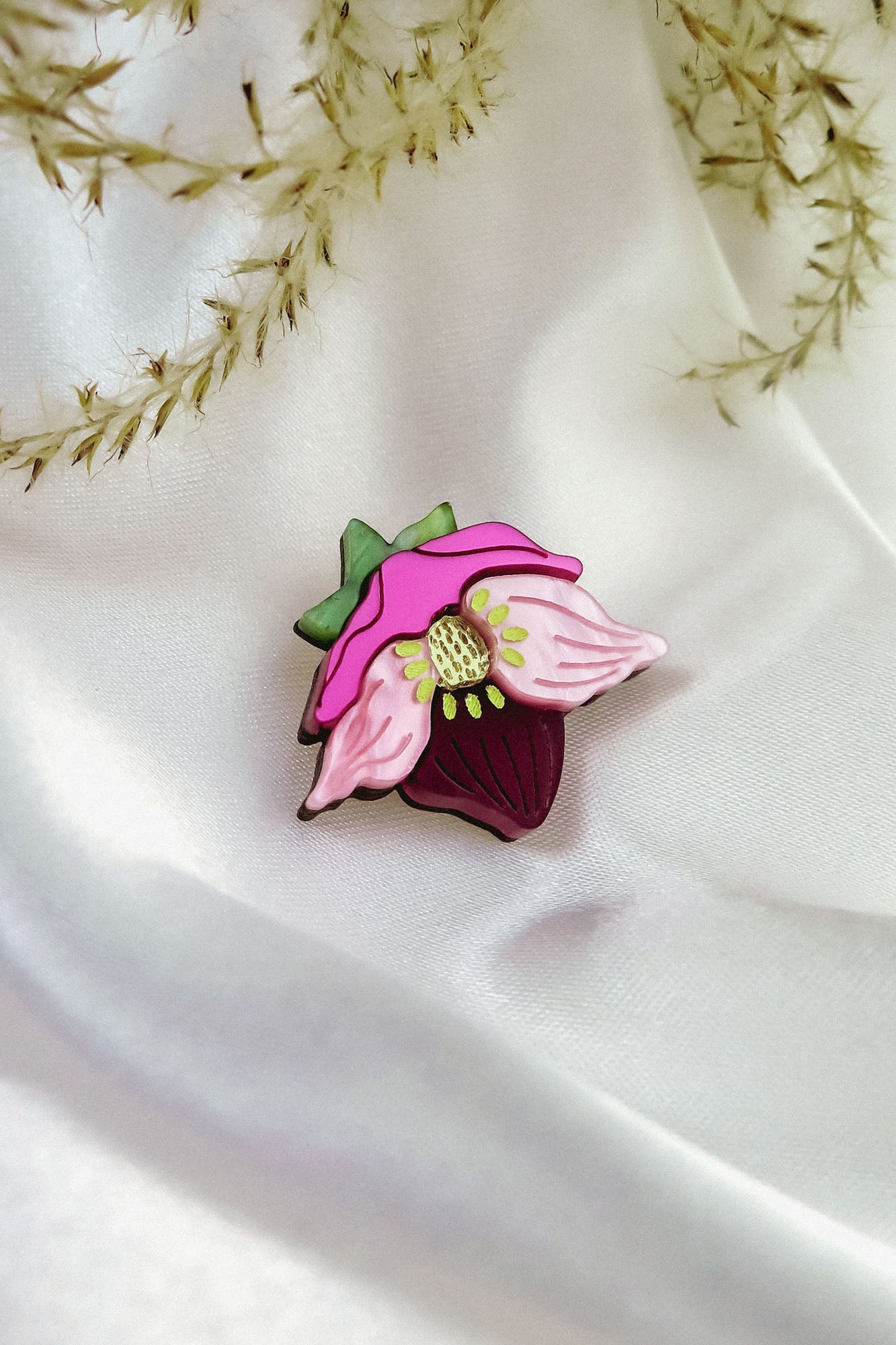A handmade Purple Hellebore Flower lapel pin rests on white satin fabric, with delicate sprigs of dried grass in the background.