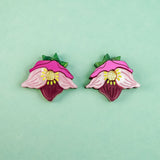 Two colorful Hellebore Stud Earrings, shaped like stylized pink flowers with green leaves and yellow centers, are displayed on a light turquoise background—perfect handcrafted floral jewelry for unique accessories lovers.