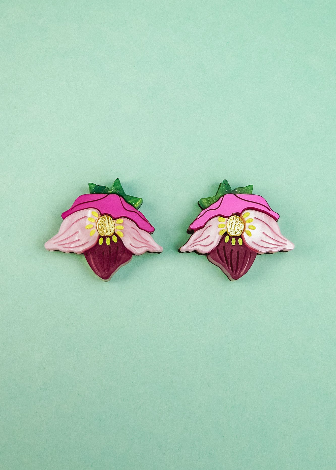 Two colorful Hellebore Stud Earrings, shaped like stylized pink flowers with green leaves and yellow centers, are displayed on a light turquoise background—perfect handcrafted floral jewelry for unique accessories lovers.