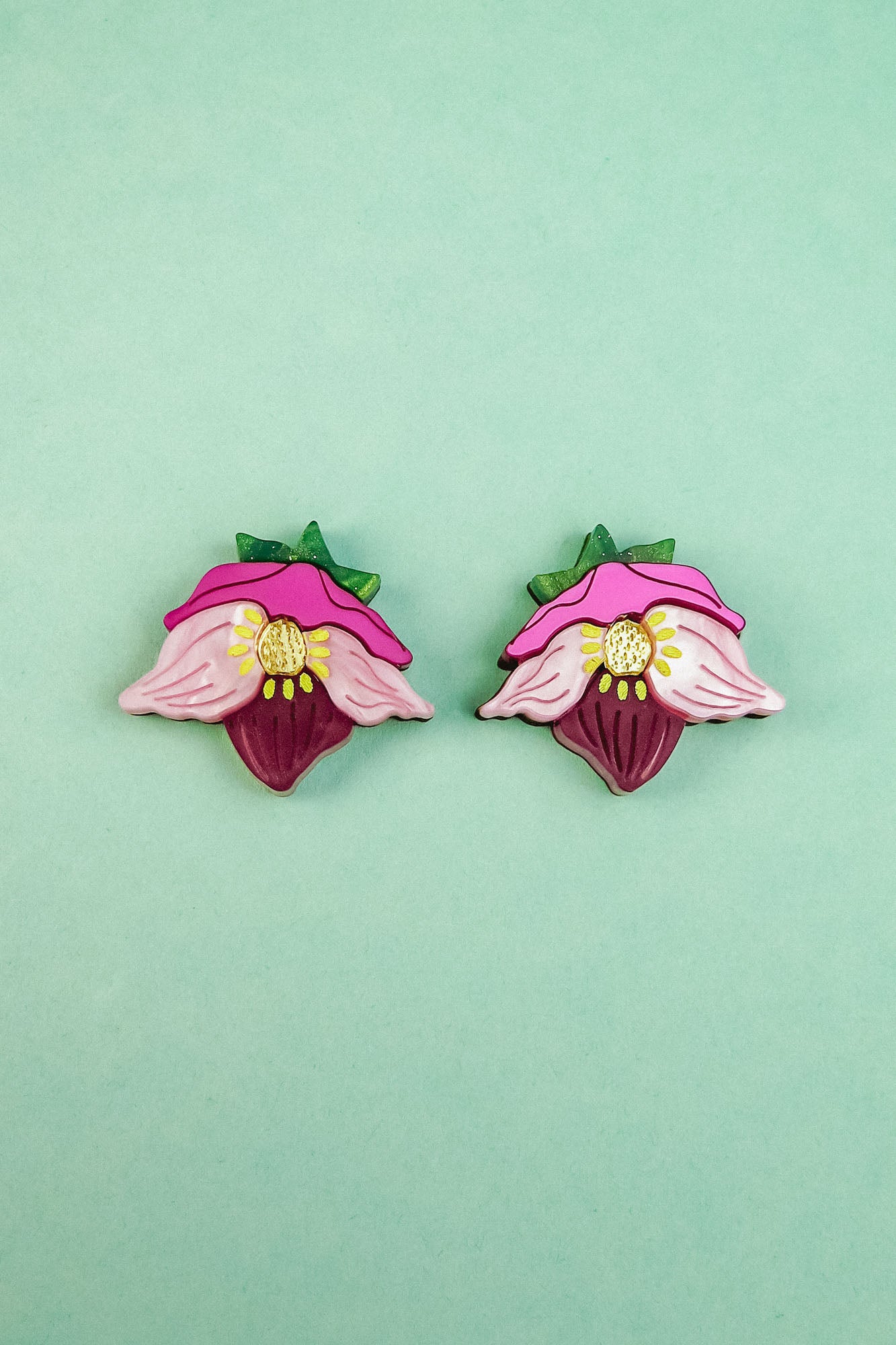 Two colorful Hellebore Stud Earrings, shaped like stylized pink flowers with green leaves and yellow centers, are displayed on a light turquoise background—perfect handcrafted floral jewelry for unique accessories lovers.