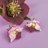 A pair of Hellebore Stud Earrings with pink, white, and green accents are displayed on a purple surface next to a real deep pink flower with yellow stamens and green leaves—perfect handcrafted floral jewelry for unique accessories lovers.