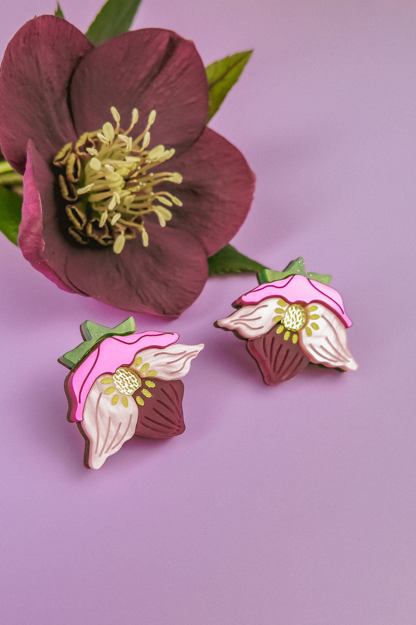 A pair of Hellebore Stud Earrings with pink, white, and green accents are displayed on a purple surface next to a real deep pink flower with yellow stamens and green leaves—perfect handcrafted floral jewelry for unique accessories lovers.