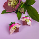 Two cupcake-shaped Hellebore Stud Earrings with pink and white flower designs are displayed on a lilac background next to a purple flower with green leaves, showcasing handcrafted floral jewelry for unique accessories lovers.