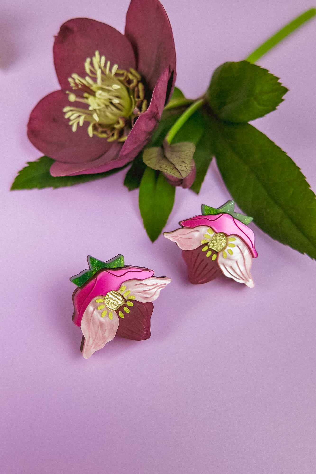 Two cupcake-shaped Hellebore Stud Earrings with pink and white flower designs are displayed on a lilac background next to a purple flower with green leaves, showcasing handcrafted floral jewelry for unique accessories lovers.