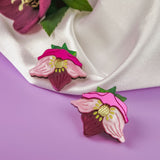 Two decorative pins shaped like strawberries with pink flower petals and green leaves rest on a light purple surface next to a white satin fabric and a dark pink flower, perfect for fans of unique accessories and handcrafted floral jewelry.