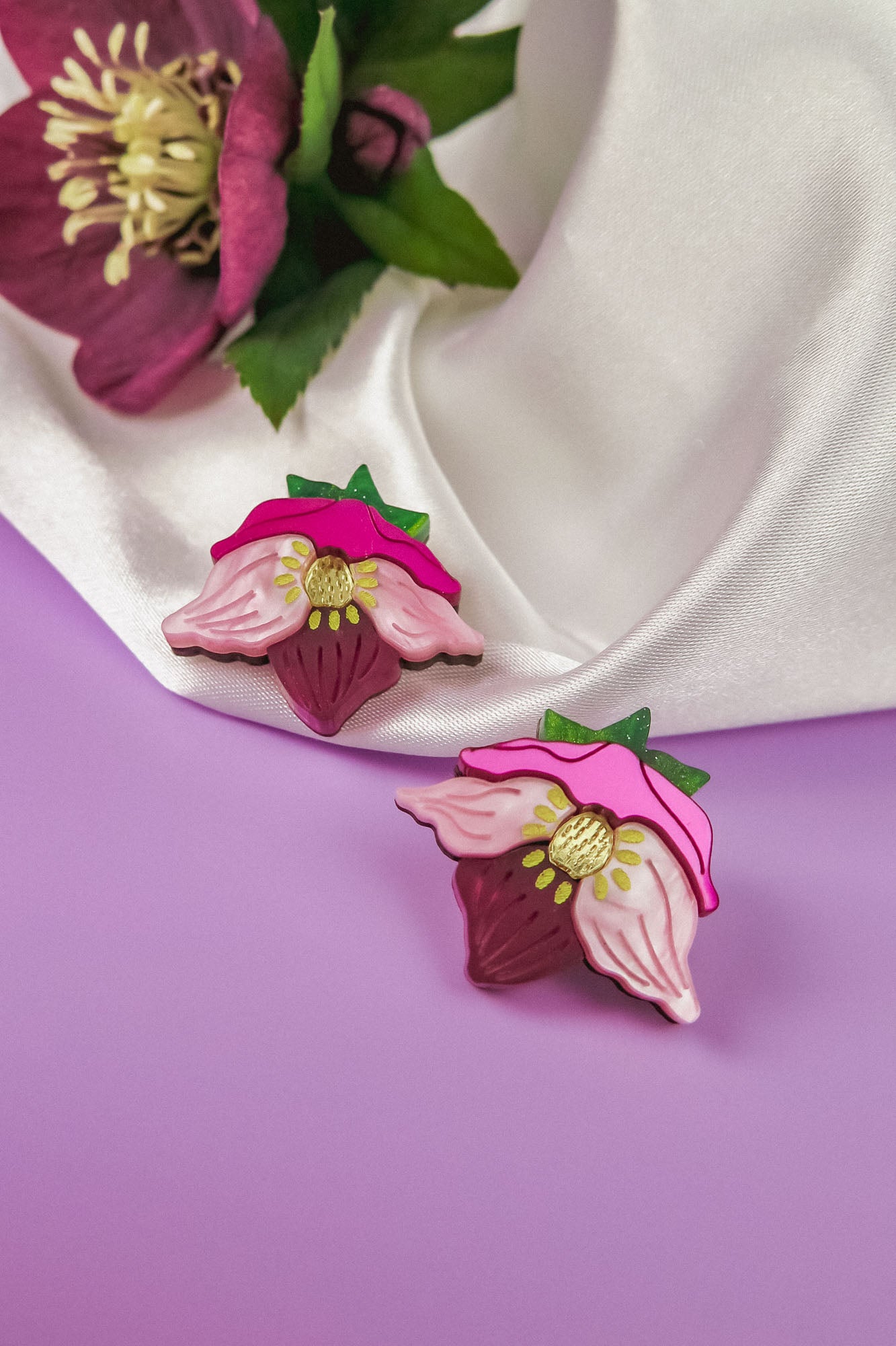 Two decorative pins shaped like strawberries with pink flower petals and green leaves rest on a light purple surface next to a white satin fabric and a dark pink flower, perfect for fans of unique accessories and handcrafted floral jewelry.