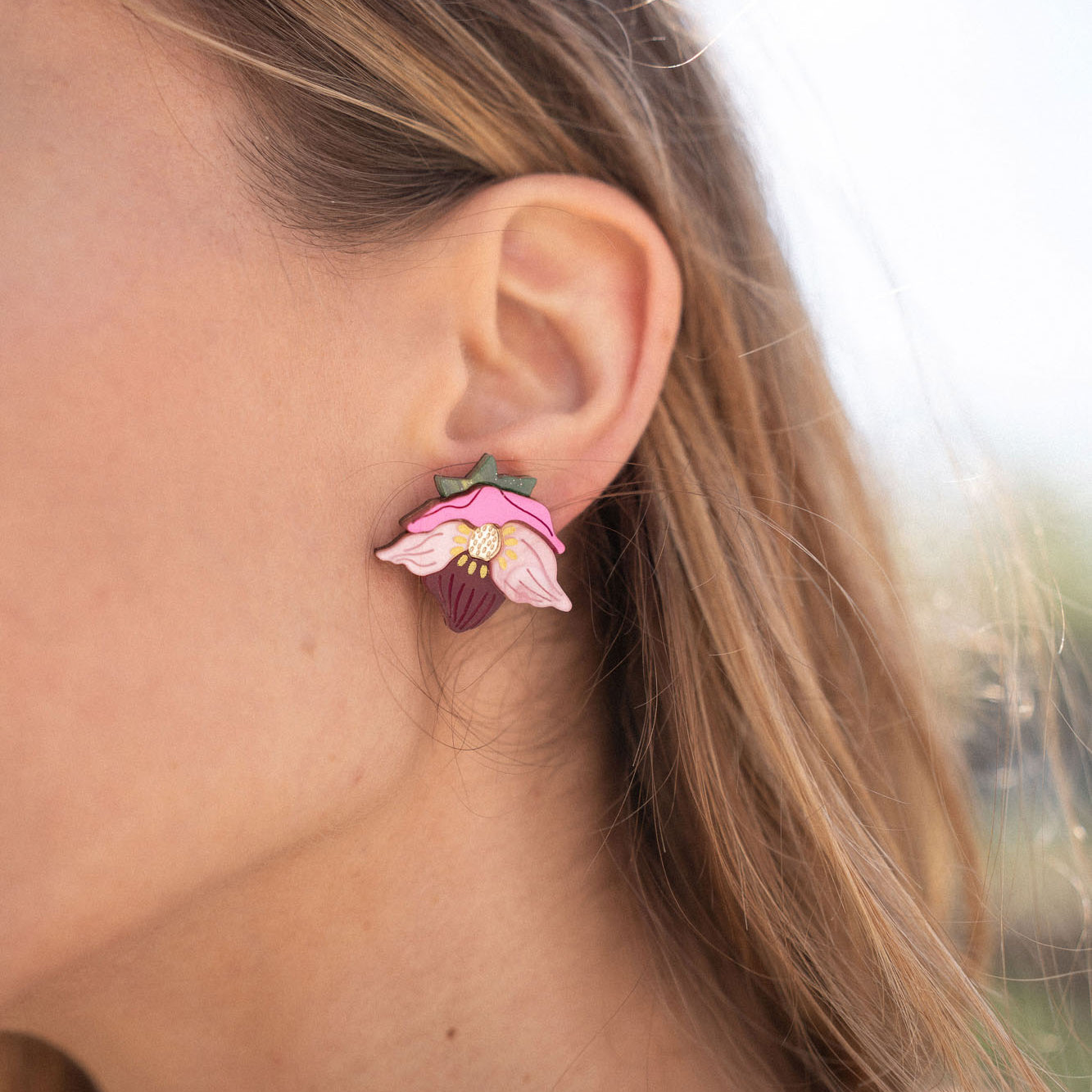 A close-up of a person’s ear wearing Hellebore Stud Earrings, handcrafted floral jewelry with pink petals and a purple center. The person has long light brown hair and is outdoors in natural light, showcasing these unique accessories.