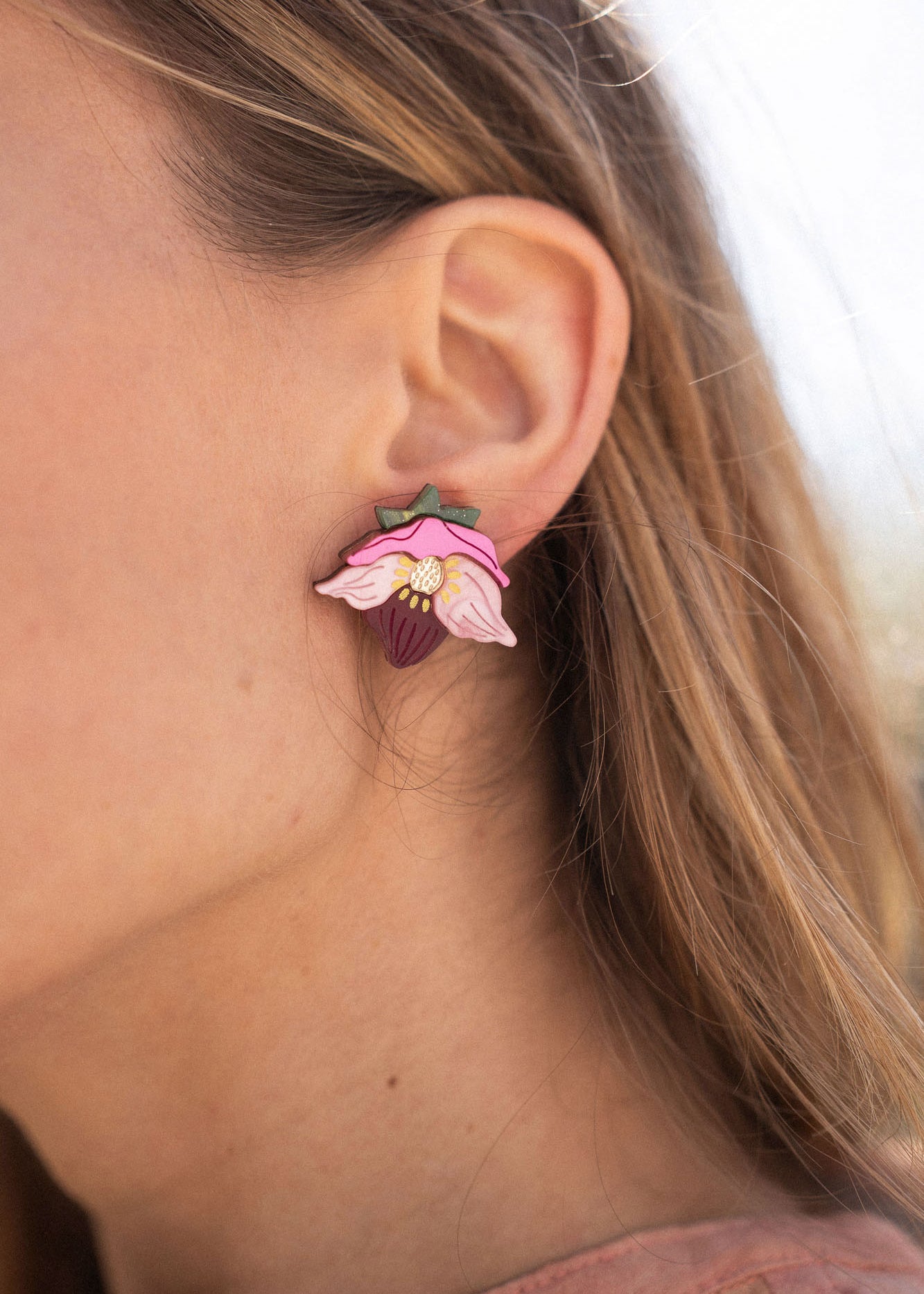 A close-up of a person’s ear wearing Hellebore Stud Earrings, handcrafted floral jewelry with pink petals and a purple center. The person has long light brown hair and is outdoors in natural light, showcasing these unique accessories.