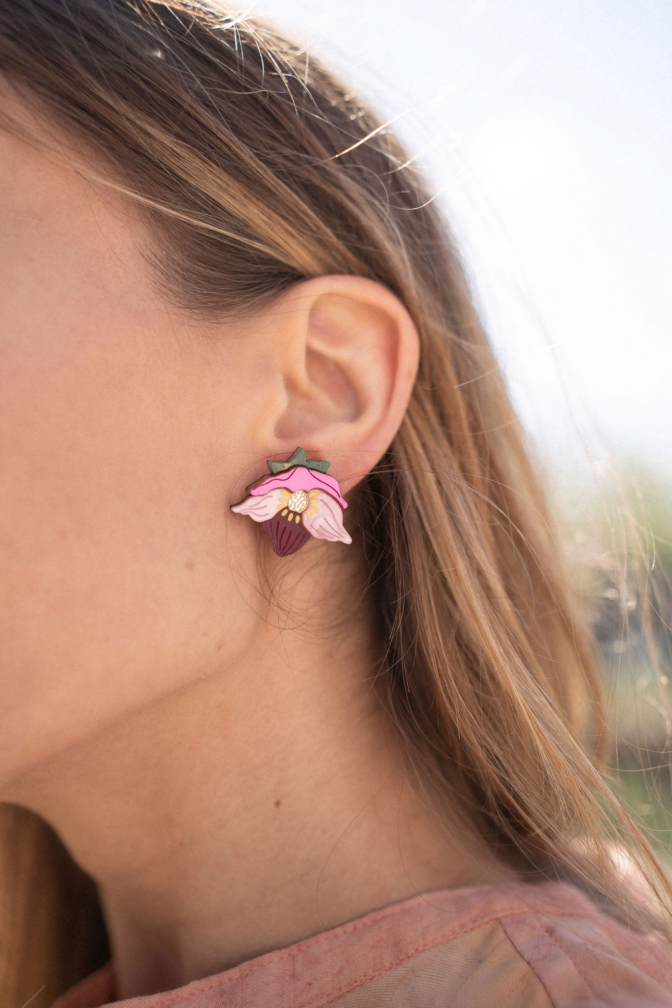 A close-up of a person’s ear wearing Hellebore Stud Earrings, handcrafted floral jewelry with pink petals and a purple center. The person has long light brown hair and is outdoors in natural light, showcasing these unique accessories.