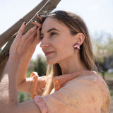 A woman with long hair wearing a peach-colored blouse and handcrafted floral jewelry stands outdoors, gently touching a tree branch and smiling softly in natural sunlight.