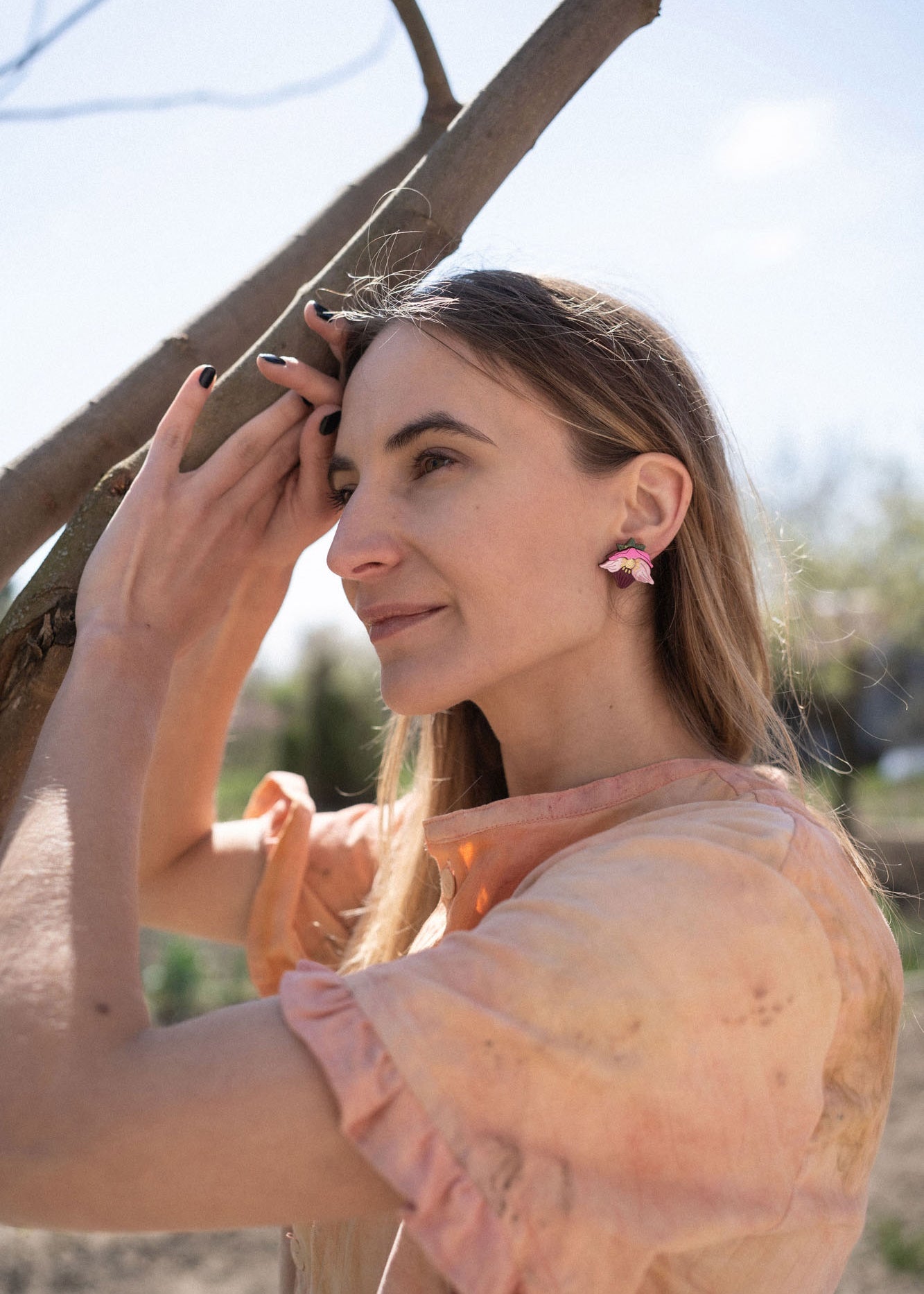 A woman with long hair wearing a peach-colored blouse and handcrafted floral jewelry stands outdoors, gently touching a tree branch and smiling softly in natural sunlight.