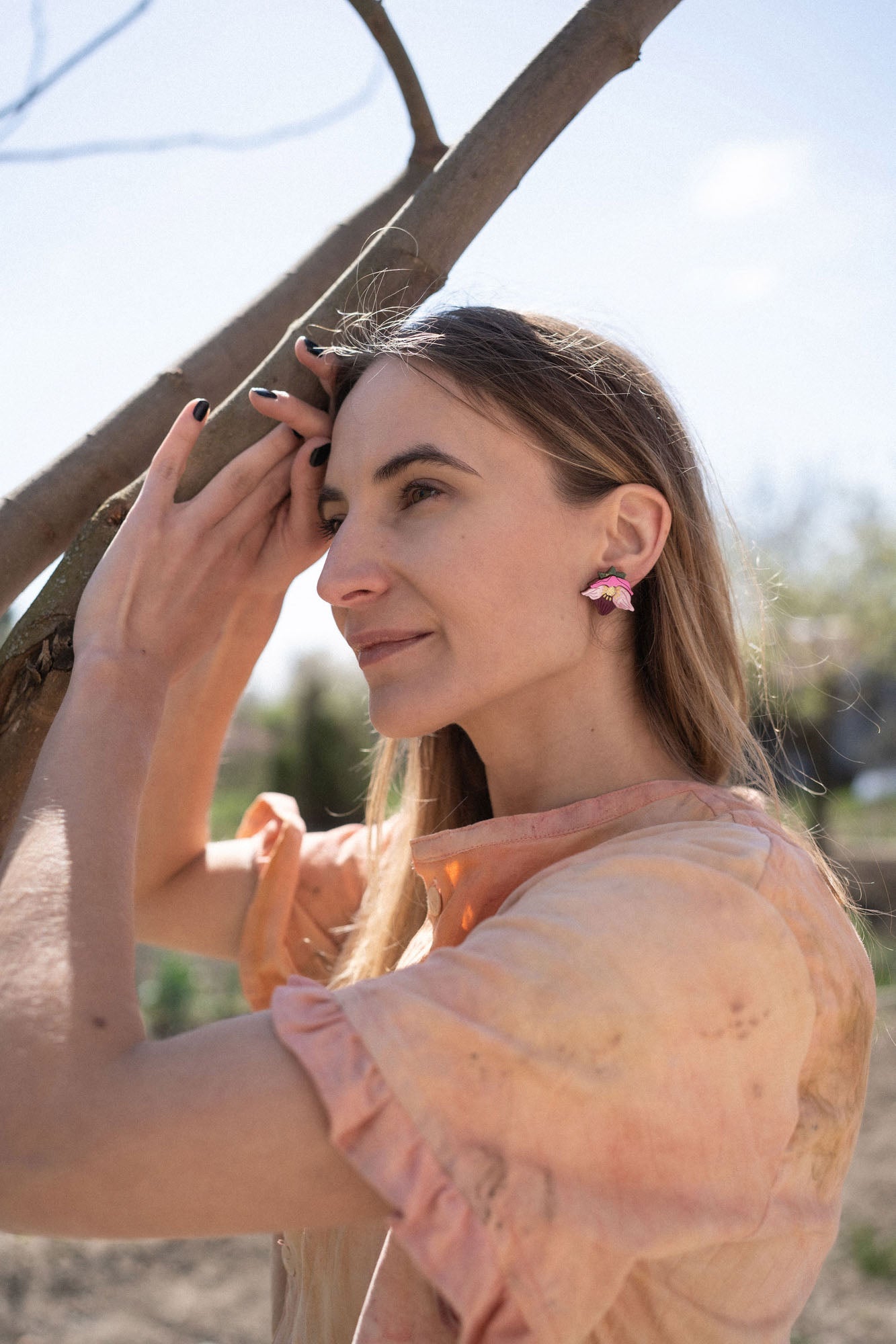 A woman with long hair wearing a peach-colored blouse and handcrafted floral jewelry stands outdoors, gently touching a tree branch and smiling softly in natural sunlight.
