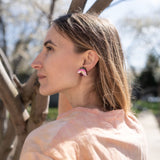 A woman with light brown hair stands outdoors near tree branches, wearing a light pink shirt and unique accessories—colorful, handcrafted floral jewelry that includes striking pink Hellebore Stud Earrings—as she gazes thoughtfully into the distance on a sunny day.