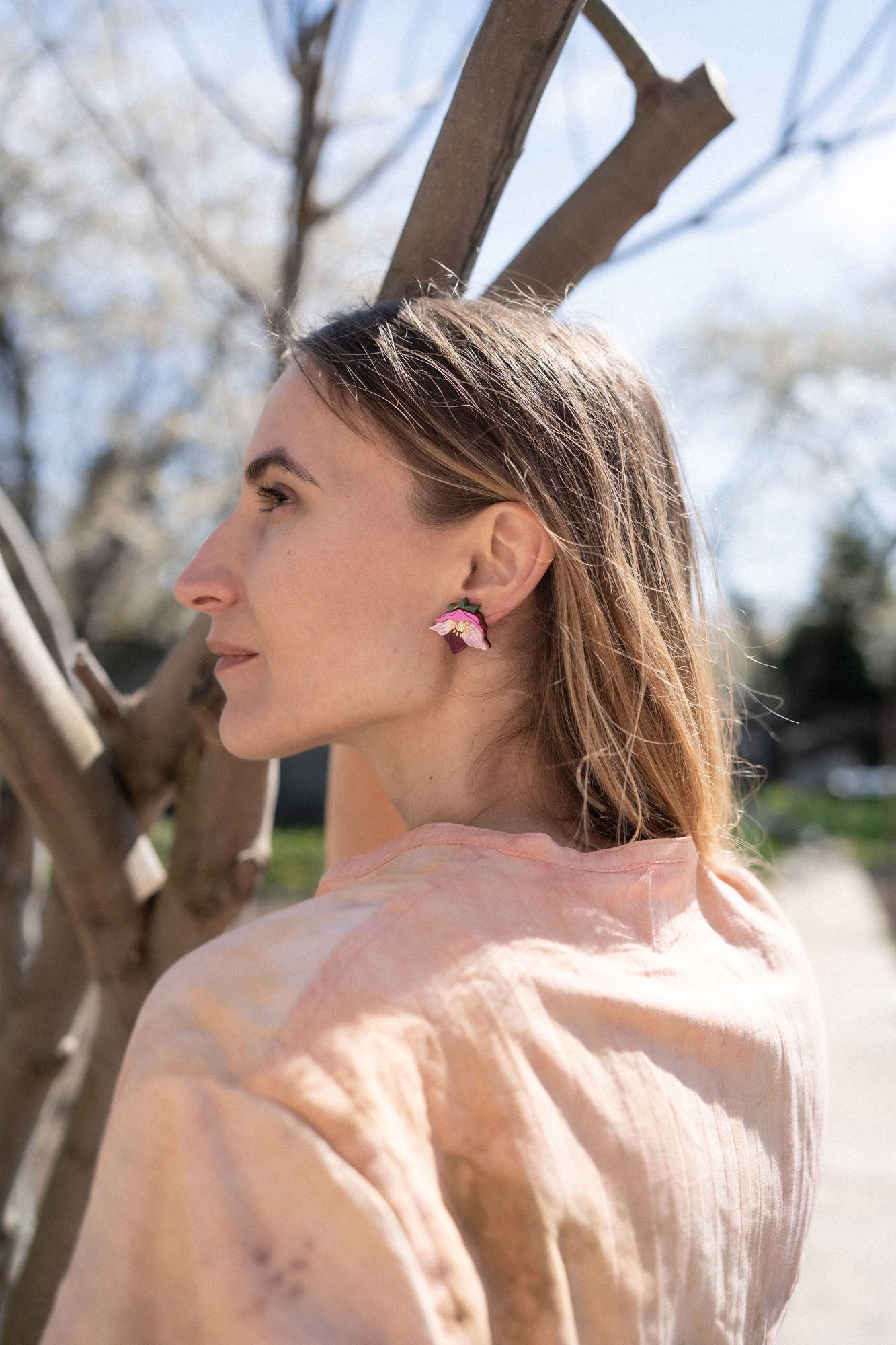 A woman with light brown hair stands outdoors near tree branches, wearing a light pink shirt and unique accessories—colorful, handcrafted floral jewelry that includes striking pink Hellebore Stud Earrings—as she gazes thoughtfully into the distance on a sunny day.