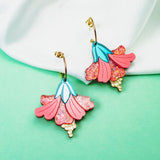 A pair of vibrant Hibiscus Earrings with gold hooks rest on a soft white fabric against a mint green background. These tropical statement earrings feature pink petals, glittery accents, blue details, and small yellow bead-like clusters.