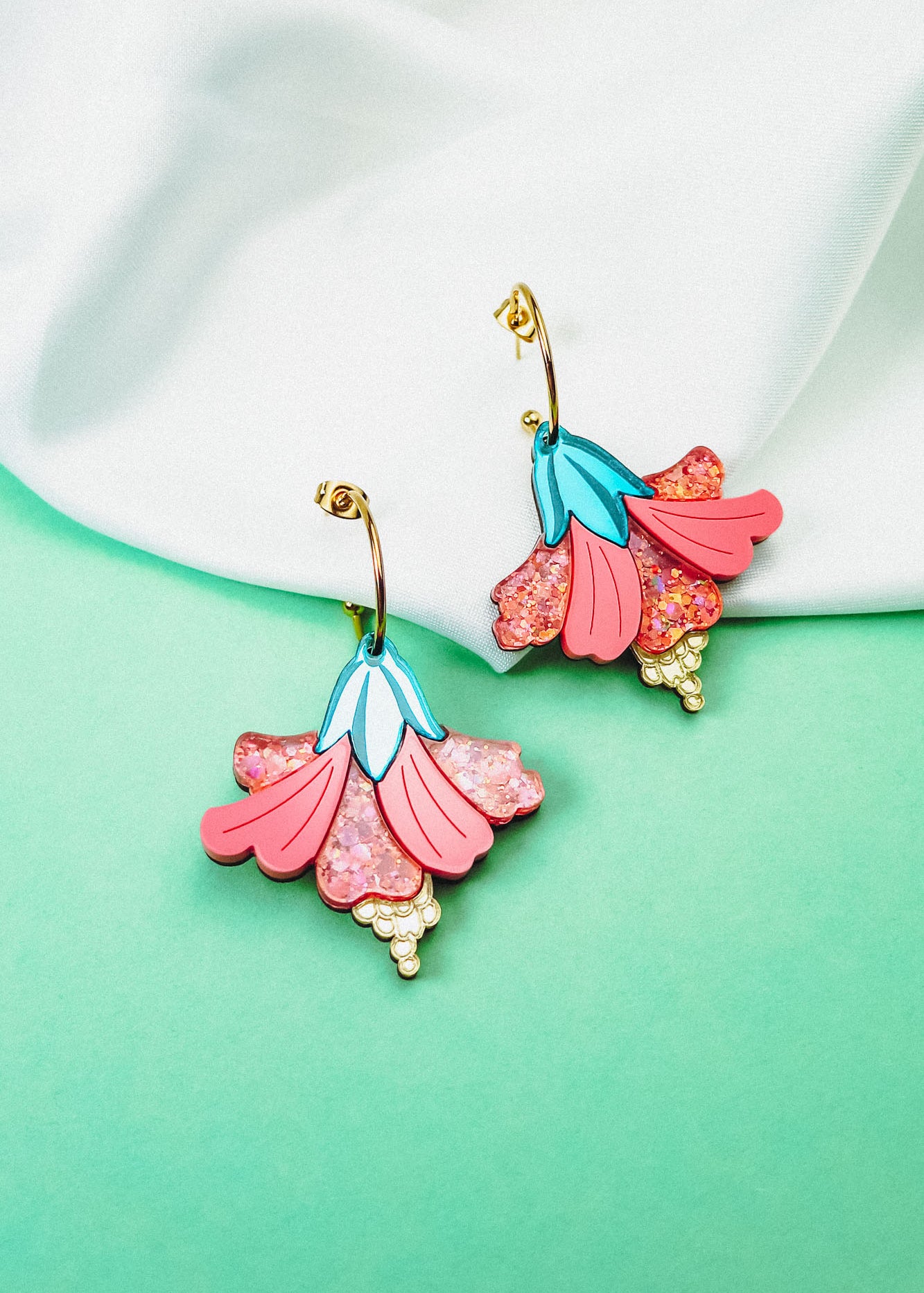 A pair of vibrant Hibiscus Earrings with gold hooks rest on a soft white fabric against a mint green background. These tropical statement earrings feature pink petals, glittery accents, blue details, and small yellow bead-like clusters.