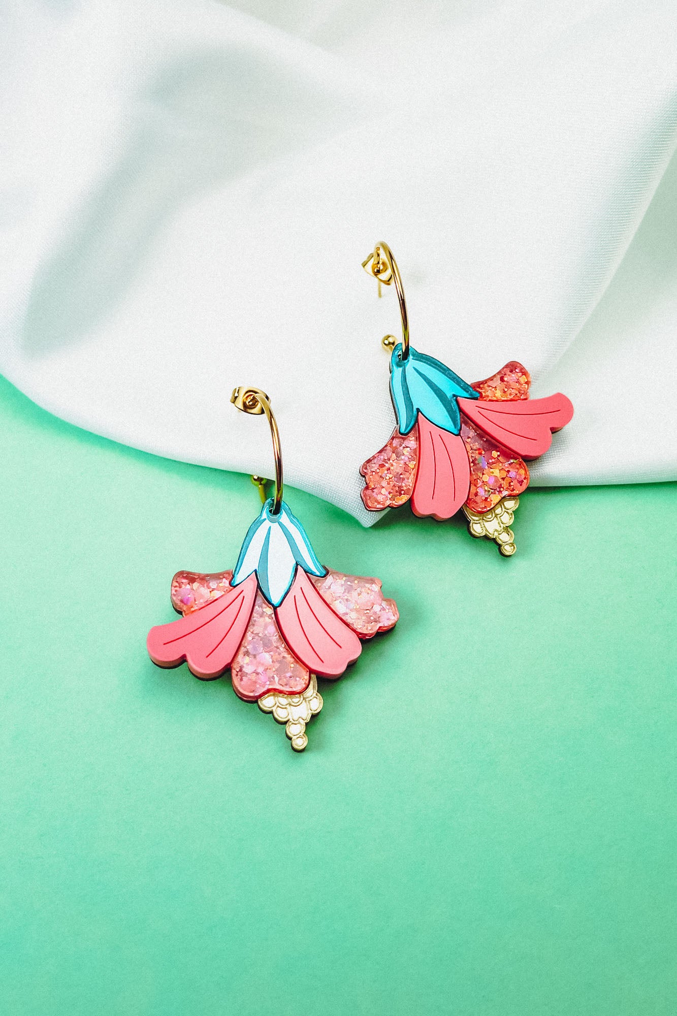 A pair of vibrant Hibiscus Earrings with gold hooks rest on a soft white fabric against a mint green background. These tropical statement earrings feature pink petals, glittery accents, blue details, and small yellow bead-like clusters.