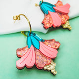 Vibrant Hibiscus Earrings with gold hooks, featuring colorful pink, blue, and glittery accents, make a bold tropical statement against a striking green and white background.
