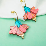 A vibrant pair of Hibiscus Earrings with gold hooks, featuring pink and blue petals and sparkling pink accents, make a tropical statement against a white cloth and mint green background.